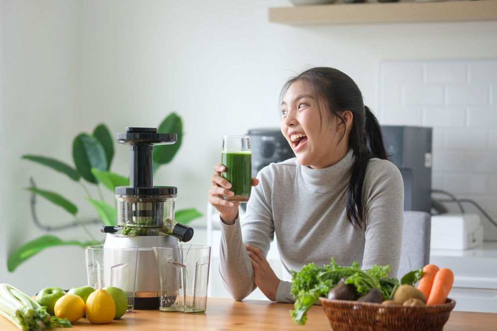 Cheerful woman enjoying a glass of fresh cold-pressed green vegetable juice in a bright kitchen. Healthy lifestyle and wellness concept