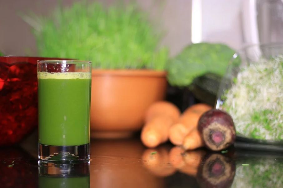 a glass of green juice with fruits and vegetables in the background