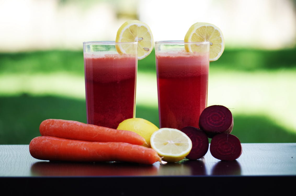two glasses of juices in front of carrots, lemons, and beets